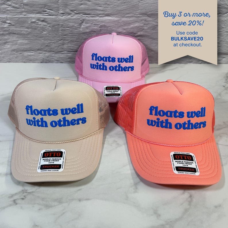 May include: Three trucker hats in light pink, beige, and coral, each featuring a mesh back and the blue text "floats well with others" on the front foam panel. Each hat includes an Otto brand sticker on the brim and a snapback closure.