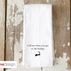 May include: White kitchen towel with the text "Call me when you get to the bridge." and a black graphic of a coastline. The towel is folded and rests on a weathered wooden surface.