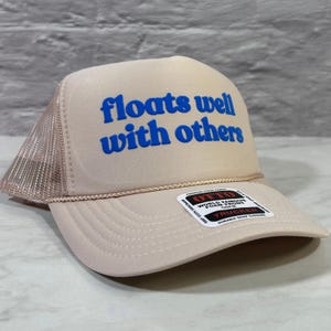 May include: A beige trucker hat featuring a mesh back and a foam front panel with the blue text "floats well with others" printed in a bold, rounded font. A rectangular sticker with the brand name Otto is attached to the brim of the hat.