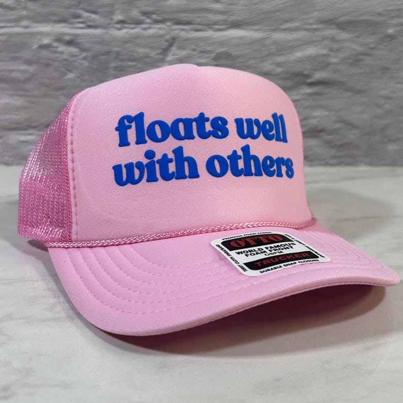 May include: A light pink trucker hat featuring a mesh back and a foam front panel. The text "floats well with others" is printed in bold blue letters on the front. A black and white rectangular sticker is attached to the brim of the hat.