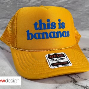 May include: Yellow trucker hat with the phrase "this is bananas" in blue lettering. The hat has a mesh back and a snap closure. A small label is visible on the front.