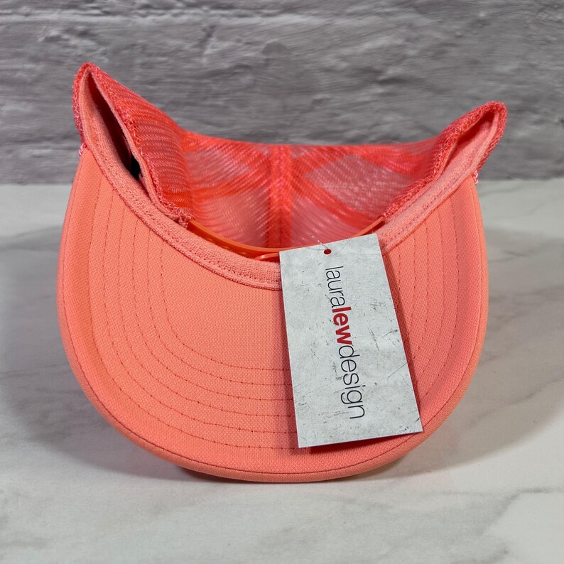 May include: A bright coral trucker hat featuring a mesh back panel and a solid front brim. A rectangular white paper tag is attached to the brim, displaying the text lauralewdesign in black and red lettering, with a distressed texture.