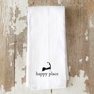 May include: A white rectangular kitchen towel featuring a black graphic of the Cape Cod peninsula silhouette positioned above the lowercase black text that reads happy place centered near the bottom edge of the fabric.