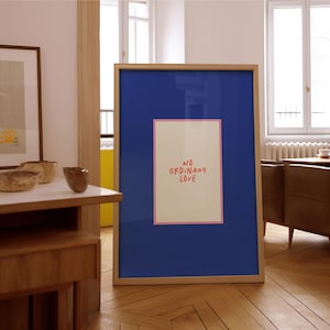 May include: A framed art print with a wooden frame. The artwork features a blue background with a rectangular off-white centre, outlined in pink, with the words "NO ORDINARY LOVE" in red. The print is displayed on a wooden floor.