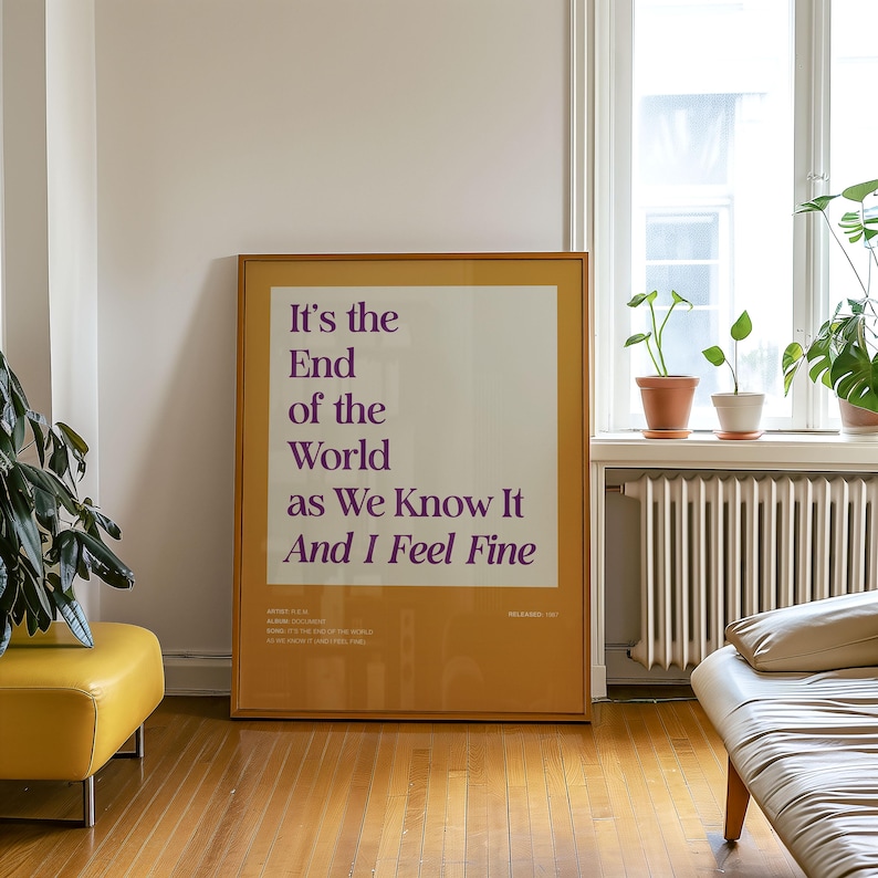 Puede incluir: Un p&oacute;ster enmarcado con el texto "It's the End of the World as We Know It And I Feel Fine" en morado. El p&oacute;ster tiene un marco dorado y est&aacute; apoyado contra una pared blanca. La habitaci&oacute;n tiene un otomana amarilla y una ventana con plantas.