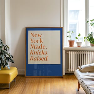 May include: A rectangular framed wall art piece featuring a blue border and a cream-colored center panel. The text "New York Made. Knicks Raised." is printed in a bold, orange serif font centered within the cream-colored rectangular area.