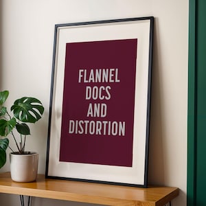 May include: Framed art print with the words "FLANNEL DOCS AND DISTORTION" in white on a burgundy background. The print is in a black frame and sits on a wooden surface. A potted plant with large green leaves is to the left.