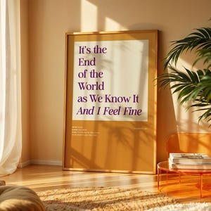 Puede incluir: Un p&oacute;ster enmarcado con el texto "It's the End of the World as We Know It And I Feel Fine" en morado sobre un fondo crema. El p&oacute;ster est&aacute; en una habitaci&oacute;n con una ventana, una planta y una peque&ntilde;a mesa naranja.