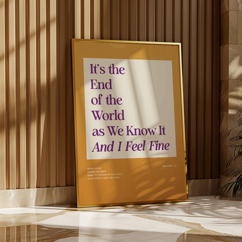 Puede incluir: Un p&oacute;ster enmarcado con el texto "It's the End of the World as We Know It And I Feel Fine" en morado sobre un fondo crema. El p&oacute;ster est&aacute; en un marco dorado y se apoya en una pared de madera.
