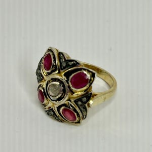 May include: A gold ring with a floral design. The ring features five red gemstones and a central clear gemstone. The setting is accented with small black stones. The ring is set against a white background.