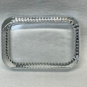 May include: A clear, rectangular glass tray with rounded corners. The edges are adorned with a series of small, evenly spaced, raised bumps. The tray is set against a white background.