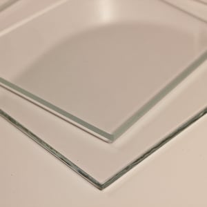 May include: Two clear, rectangular glass panels are stacked at a slight angle, showcasing their transparency. The edges of the glass are visible, and the panels are resting on a light-colored surface. The image highlights the clarity and shape of the glass.