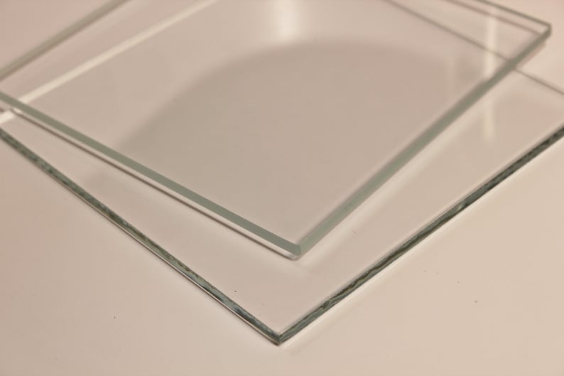 May include: Two clear, rectangular glass panels are stacked at a slight angle, showcasing their transparency. The edges of the glass are visible, and the panels are resting on a light-colored surface. The image highlights the clarity and shape of the glass.