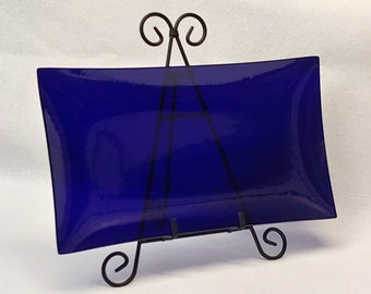 May include: A cobalt blue glass serving tray with a curved edge, displayed on a black metal stand with decorative scrolls.