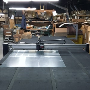 May include: A large industrial glass cutting machine with a clear glass sheet in place. The machine has a black and silver frame, with a cutting head poised above the glass. The background shows a cluttered workshop with shelves and various materials.