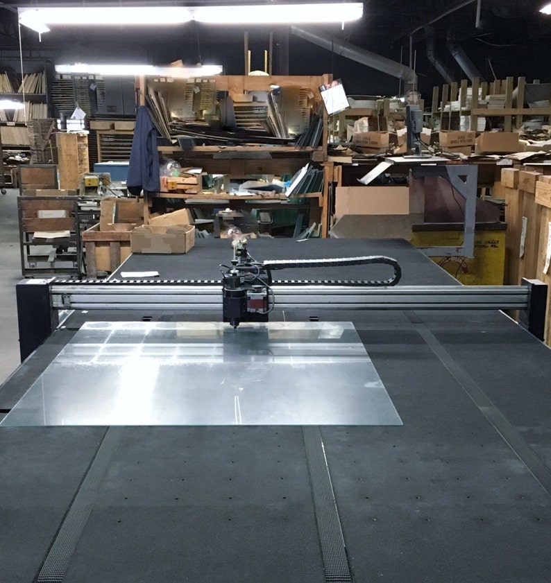May include: A large industrial glass cutting machine with a clear glass sheet in place. The machine has a black and silver frame, with a cutting head poised above the glass. The background shows a cluttered workshop with shelves and various materials.