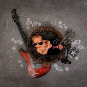 May include: A newborn baby wearing sunglasses is lying in a wooden bowl with a red electric guitar, a microphone, and several ticket stubs scattered around the bowl.