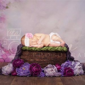 May include: A sleeping newborn baby girl wearing a pink headband with a flower on top, lying in a brown wicker basket lined with green fabric, surrounded by purple flowers on a wooden floor.