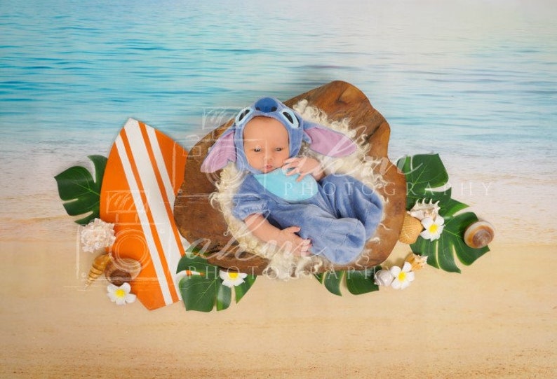 Puede incluir: Un beb&eacute; con un disfraz azul de Stitch est&aacute; acostado en un cuenco de madera con una tabla de surf y hojas tropicales sobre un fondo de playa.