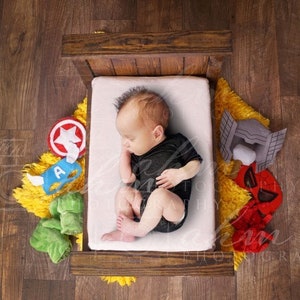 May include: A newborn baby sleeping in a wooden crib with a white sheet. The crib is surrounded by superhero-themed toys, including a red and yellow star, a blue and yellow mask, a green and yellow Hulk hand, a red and black Spider-Man mask, and a gray and silver Thor helmet.