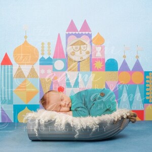 May include: A newborn baby sleeping in a gray boat-shaped bassinet with a white fluffy lining. The baby is wearing a teal blue onesie. The background is a blue wall with a colorful illustration of a whimsical castle.