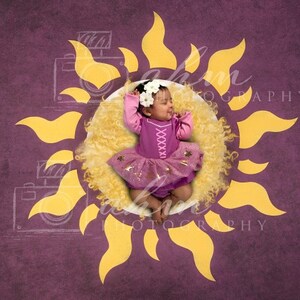 May include: A baby dressed in a purple and gold princess costume is lying on a yellow fluffy blanket inside a yellow sun cutout. The background is a deep purple.