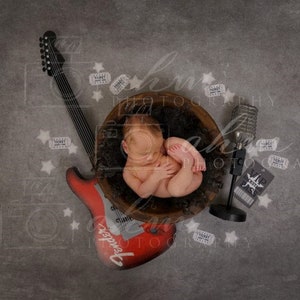 May include: A newborn baby is sleeping in a wooden bowl with a red electric guitar, a microphone, and several tickets scattered around. The guitar has the word "Fender" written on it.