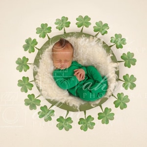 May include: A newborn baby wearing a green outfit is sleeping in a white fluffy nest surrounded by a circle of green shamrock leaves.
