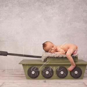 May include: A newborn baby sleeping on a green toy tank with a brown barrel. The tank has four wheels and is on a wooden floor.