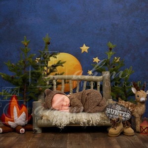 May include: A newborn baby sleeping on a rustic wooden bench with a faux fur blanket. The bench is decorated with a sign that says "Little Hunter in Training". There is a stuffed deer and a campfire behind the bench.