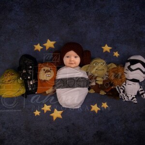 May include: A baby wrapped in a white blanket with a gray pattern is surrounded by Star Wars characters. The baby is wearing a brown hat with a white trim. The characters include Yoda, Chewbacca, Darth Vader, and a Stormtrooper. The background is a dark blue with yellow stars.