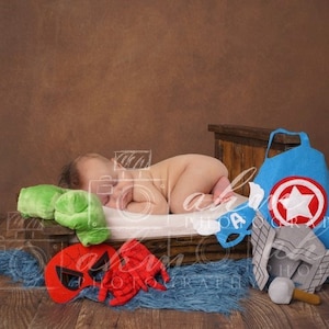 Può includere: Un neonato che dorme su un letto di legno con un mantello di supereroe blu e rosso, un pugno verde, una maschera di Spider-Man rossa e nera e un elmetto grigio con un martello.