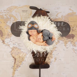 May include: A newborn baby in a gray aviator hat and pants, nestled in a fluffy white nest, posed on a wooden airplane prop. The backdrop is a world map. The airplane prop has a propeller and wings.