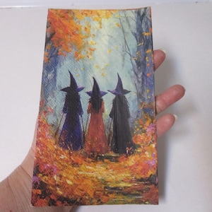 May include: A rectangular paper napkin featuring a printed illustration of three figures wearing pointed hats and long robes standing in a forest. The scene depicts a path covered in vibrant orange and yellow autumn leaves with trees in the background.