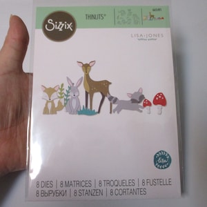 May include: A package of 8 die-cut metal shapes for crafting. The shapes include a deer, fox, rabbit, raccoon, and mushrooms. The package is labeled "Sizzix Thinlits" and "Lisa Jones" with the text "8 Dies | 8 Matrices | 8 Troqueles | 8 Fustelle | 8 Вырубки | 8 Stanzen | 8 Cortantes".