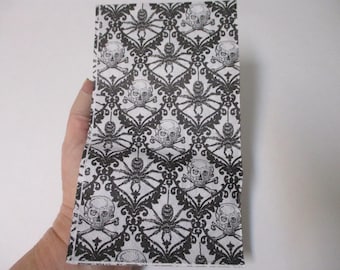 May include: A rectangular, folded item with a repeating pattern of skulls and crossbones, and spiders in black and white. The design has a damask-like quality, creating a gothic or Halloween theme. The item is held in a hand.