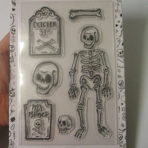 May include: Clear Halloween-themed stamp set featuring a skeleton, skulls, and tombstones. One tombstone reads "October 31st" with crossed bones. Another reads "Mr. Bones" with a skull and crossbones. The stamps are perfect for crafting.