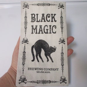 Pode incluir: Um descanso de copo de tecido branco com texto preto e uma ilustração de um gato preto. O texto diz "Black Magic Brewing Company Salem, Mass."