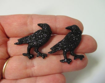 May include: Two black glitter crow figurines. The crows are standing on their feet with their wings spread out.