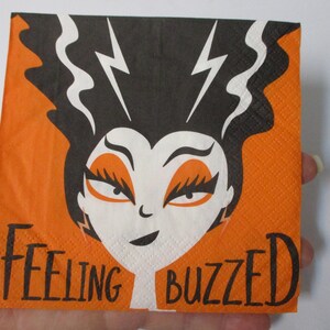May include: Orange and black paper napkin with a cartoon illustration of a female monster with black hair and lightning bolts. The text "Feeling Buzzed" is printed on the napkin.