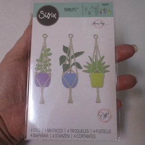 May include: A clear plastic package containing Sizzix Thinlits dies. The dies are shaped like hanging planters with colorful plants. The package includes the text "Olivia Raye" and "4 DIES".