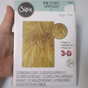 May include: A gold-colored 3-D embossing folder with a palm leaf design. The folder is packaged in a clear plastic sleeve with a white label that reads "3-D Textured Impressions" and "Georgie Cany".
