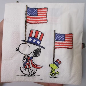 May include: A white paper napkin with a cartoon image of Snoopy and Woodstock dressed in patriotic attire. Snoopy is wearing a red, white, and blue top hat and holding a small American flag. Woodstock is wearing a red, white, and blue hat and holding a larger American flag. The napkin has the text "©PNTS" in the bottom right corner.