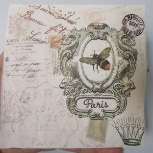 May include: A vintage-style paper napkin with a bee illustration in a decorative frame. The napkin features a faded background with handwritten text and a stamp that reads "Assilie, Touches du Rhone". The text "Paris" is written in a banner below the bee.