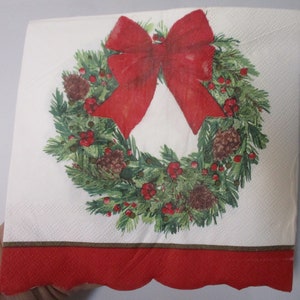 May include: A white paper napkin with a red and green Christmas wreath design. The wreath is decorated with a red bow, pine cones, and red berries.