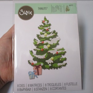 May include: A package of 8 Sizzix Thinlits dies for crafting. The dies are designed to cut out a Christmas tree with snow, ornaments, and presents. The package is white with green and brown accents. The text on the package reads "Sizzix Thinlits" and "8 Dies | 8 Matrices | 8 Troqueles | 8 Fustelle | 8 Вырубки | 8 Stanzen | 8 Cortantes".