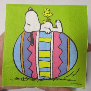 May include: Snoopy the beagle is lying on top of a large, colorful Easter egg. The egg is decorated with stripes and zigzags in shades of blue, pink, yellow, and orange. Snoopy is wearing a black nose and has a black tail.  A small yellow bird with a red beak is perched on top of Snoopy's head. The background is a solid green color. © 2022 Peanuts