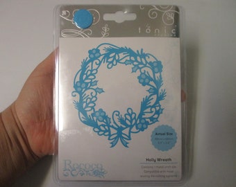 May include: A blue metal die cut of a holly wreath with a white background. The die cut is packaged in a clear plastic sleeve with a white backing card. The card includes the text "Holly Wreath", "Actual Size 80mm x 80mm 3.1" x 3.1"", and "Contains 1 metal craft die. Compatible with most leading die cutting systems."