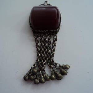 Puede incluir: Un colgante vintage con una piedra rectangular de color rojo oscuro engastada en un marco de metal plateado. El colgante presenta cadenas colgantes con pequeños dijes en forma de campana.