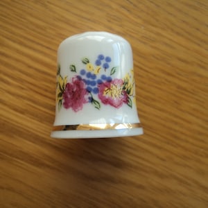 Può includere: Ditale in ceramica bianca con bordo dorato e un motivo floreale con fiori rosa, gialli e blu.